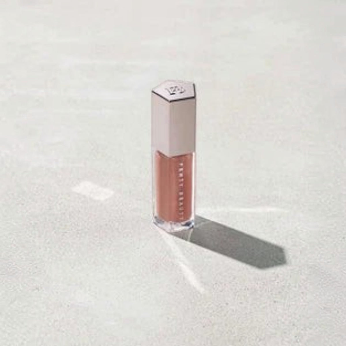 Marom Fenty Beauty Gloss Bomb Universal Lip Luminizer in a clear tube with a metallic cap on a textured surface.