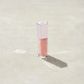Marom Fenty Beauty Gloss Bomb Universal Lip Luminizer in a clear tube with a light pink cap on a sandy surface.