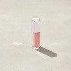 Marom Fenty Beauty Gloss Bomb Universal Lip Luminizer in a clear tube with a light pink cap on a sandy surface.
