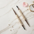 Marom ROSE BERRY dual-ended dark brown eyebrow pencil with spoolie brush on a marble surface with gold mirror